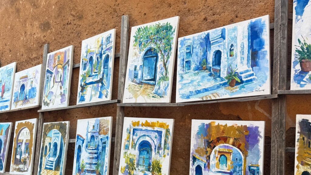 Paintings of blue alleyways hung on an ochre wall in Essaouira
