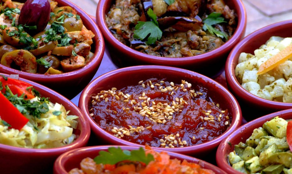 Small red bowls of Moroccan salads, including a tomato spread topped with sesame seeds, olives, aubergine and cabbage